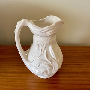 May include: A white ceramic pitcher with a large handle and a spout. The pitcher is decorated with embossed corn and wheat stalks. The pitcher is sitting on a wooden surface.