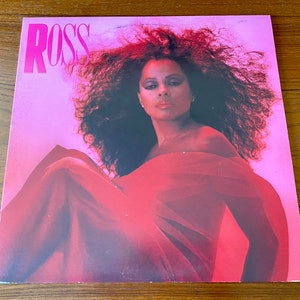 Pu&ograve; includere: Una copertina di un disco in vinile rosa con una donna con lunghi capelli scuri ricci che indossa un vestito rosso. La copertina dell'album ha la parola "ROSS" in grandi lettere rosa in alto.