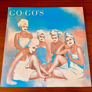 May include: Album cover for the Go-Go's "Beauty and the Beat." The image features five women in white face masks and towels, with the band's name at the top. The background is a mix of blue and pink.