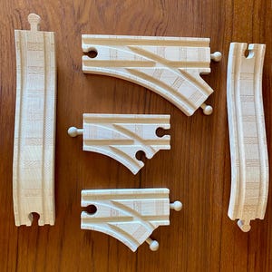 May include: Wooden toy train tracks in various shapes, including curved and straight pieces. The tracks are made of light-colored wood and feature grooves and connection points for assembling a train track layout. A classic toy for imaginative play.