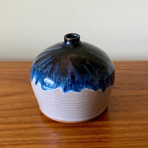 May include: A handcrafted ceramic vase with a rounded body and a small opening. The vase features a dark blue and black glazed top that transitions to a lighter blue and beige base. The vase is approximately 5 inches tall.