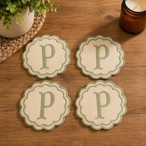 May include: A set of four round, off-white fabric coasters with scalloped edges. Each coaster features a centered, embroidered lowercase letter P in a muted sage green color, with matching green stitching along the scalloped border.
