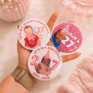 May include: Three circular pin-back buttons featuring personalized birthday designs. One button says IT'S MY WIFE'S BIRTHDAY 27, another says LAIKIN'S 27TH, and the third says IT'S MY BIRTHDAY 27 EST. 1999. Each button displays a photograph of a child and pink accents.