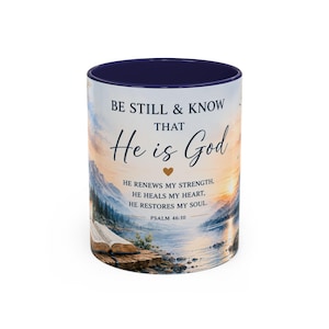 May include: A ceramic mug with a dark blue interior and rim, featuring a printed landscape of a lake, mountains, and an open book at sunset. The text reads: BE STILL & KNOW THAT He is God, HE RENEWS MY STRENGTH, HE HEALS MY HEART, HE RESTORES MY SOUL. PSALM 46:10.