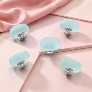 May include: Five oval-shaped cabinet knobs featuring a frosted, light blue glass top and a polished silver-tone metal base. Each knob has a smooth, rounded surface and a sturdy metallic attachment point, designed for furniture hardware applications.