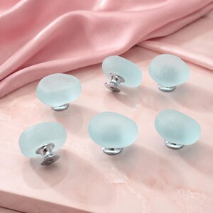 May include: A set of six cabinet knobs featuring rounded, frosted light blue glass tops with a smooth, matte texture. Each knob is attached to a polished silver-toned metal base, providing a clean, functional design for furniture hardware.
