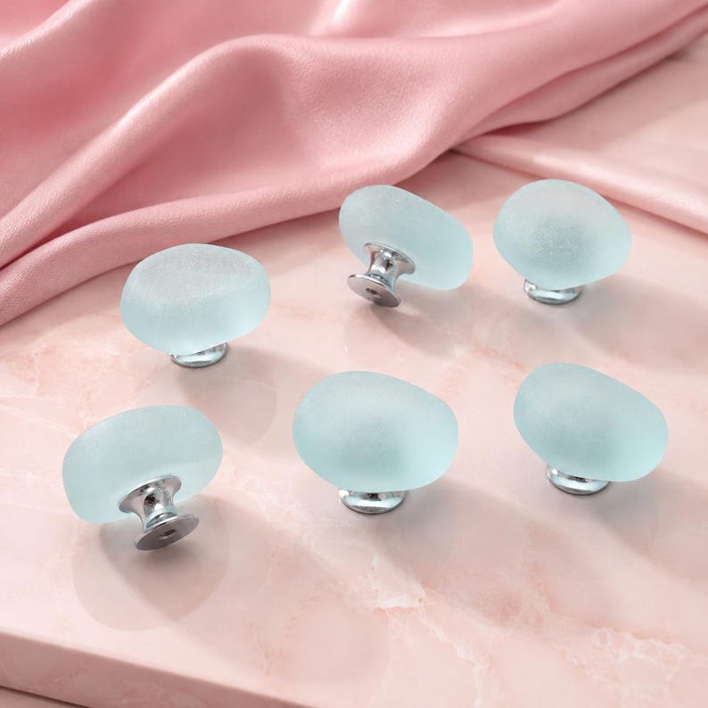 May include: A set of six cabinet knobs featuring rounded, frosted light blue glass tops with a smooth, matte texture. Each knob is attached to a polished silver-toned metal base, providing a clean, functional design for furniture hardware.