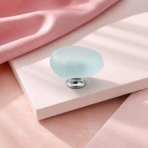 May include: A cabinet knob featuring a smooth, frosted, pale blue glass top shaped like a rounded pebble. The glass element is securely attached to a polished, circular silver-toned metal base, providing a clean and functional hardware component.