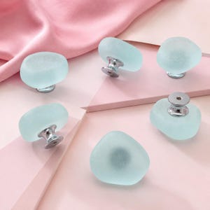 May include: Five cabinet knobs featuring smooth, frosted light blue glass tops with irregular, rounded shapes. Each knob is attached to a polished silver-tone metal base, with one knob shown from the underside revealing a threaded metal insert.