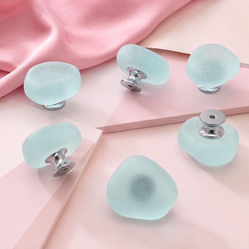 May include: Five cabinet knobs featuring smooth, frosted light blue glass tops with irregular, rounded shapes. Each knob is attached to a polished silver-tone metal base, with one knob shown from the underside revealing a threaded metal insert.