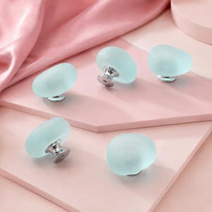 May include: Five cabinet knobs featuring smooth, frosted light blue glass tops with a rounded, pebble-like shape. Each knob is attached to a polished silver-tone metal base, providing a clean and simple look for furniture hardware applications.