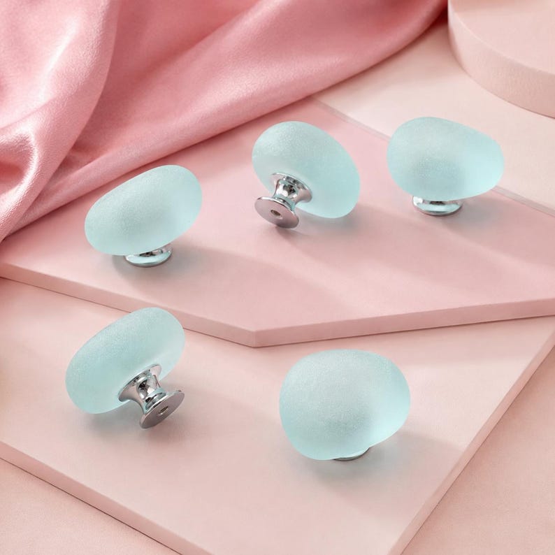 May include: Five cabinet knobs featuring smooth, frosted light blue glass tops with a rounded, pebble-like shape. Each knob is attached to a polished silver-tone metal base, providing a clean and simple look for furniture hardware applications.