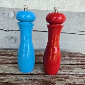 May include: Two tall, hourglass-shaped pepper mills with a glossy finish. One mill is bright blue and the other is vibrant red. Both feature a rounded top section and a metallic silver-colored adjustment knob at the very peak of each unit.