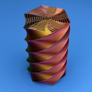 May include: A cylindrical object with a hexagonal top and a spiraling, ribbed exterior. The surface features a gradient color transition shifting between metallic gold, copper, and deep magenta tones, creating a textured, layered appearance.
