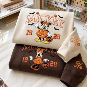 May include: A cream-colored sweatshirt featuring an embroidered Mickey Mouse in a pumpkin costume with the text MICKEY 1928, and a dark brown sweatshirt featuring an embroidered Minnie Mouse in a pumpkin costume with the text MINNIE 1928.