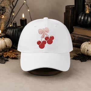 May include: A white baseball cap featuring an embroidered graphic of two red cherry-shaped silhouettes resembling mouse ears, connected by a light pink bow. The cap has a structured crown, a curved brim, and a button at the top centre.