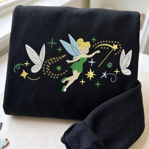 Op de afbeelding: Een zwart sweatshirt met een borduursel van een fee met blond haar, een groene jurk en blauwe vleugels. Het ontwerp bevat gele sterren, een toverstaf en extra witte vleugels met groene accenten aan de zijkanten van de afbeelding.