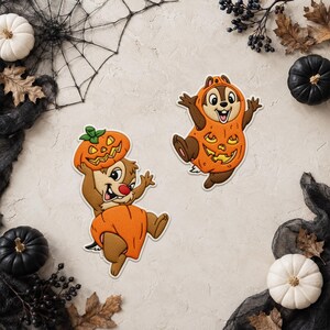 Peut inclure: Deux écussons brodés représentant des tamias marron en costumes d'Halloween. Un tamia porte un costume de citrouille et tient une citrouille d'Halloween sur sa tête. L'autre tamia porte un costume intégral de citrouille orange.