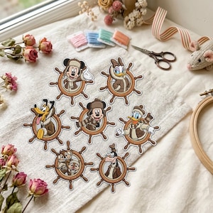 May include: A collection of seven embroidered patches featuring Disney characters including Mickey Mouse, Minnie Mouse, Donald Duck, Daisy Duck, Pluto, Goofy, and Chip and Dale, each framed within a brown ship's wheel design with winter clothing.