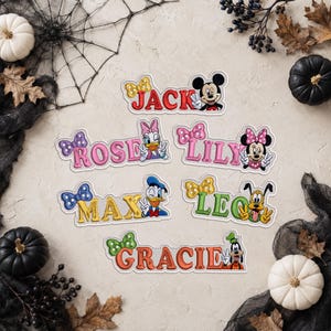 Könnte beinhalten: Sechs bestickte Namensaufnäher mit Disney-Figuren. Die Aufnäher zeigen die Namen Jack, Rose, Lily, Max, Leo und Gracie in bunten Schriftarten, jeweils begleitet von einer passenden Schleife und einer Figur wie Mickey, Daisy, Minnie, Donald, Pluto oder Goofy.