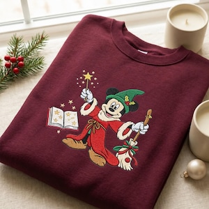 May include: A maroon crewneck sweatshirt featuring an embroidered graphic of Mickey Mouse dressed as a sorcerer in a red robe and green hat, holding a magic wand and a broom, with an open book and gold stars surrounding the character.