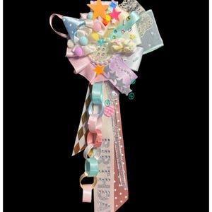 May include: A Macaron Birthday Ribbon Brooch featuring a layered arrangement of pastel ribbons, lace, and fabric stars. The accessory includes a dangling paper chain, small pom-poms, and a long ribbon tail with the word BIRTHDAY printed in silver.