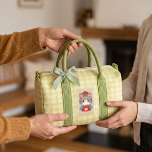 May include: A light green barrel-shaped handbag featuring a subtle diamond pattern, two green fabric handles, and a small sage green ribbon bow. A rectangular patch on the front displays an embroidered illustration of a cat wearing a red hat.