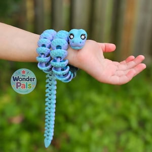 Peut inclure: Un serpent articulé et flexible présentant un dégradé de couleurs allant du bleu clair au violet. Le serpent possède un corps texturé et segmenté, une tête arrondie avec de grands yeux noirs, et est enroulé autour d'un poignet.