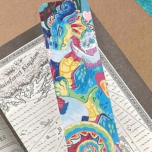 K&ouml;nnte beinhalten: Ein rechteckiges Lesezeichen mit einer farbenfrohen Illustration eines blauen und gr&uuml;nen Drachen mit einem kleinen wei&szlig;en, faultierartigen Wesen. Das Kunstwerk enth&auml;lt ein lebhaftes Spiralmuster am unteren Rand und ein kleines rosa Herz.