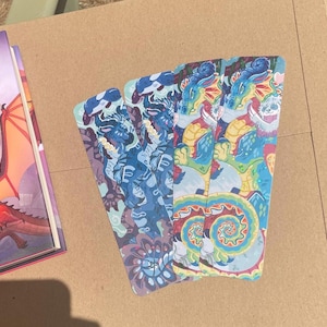 Peut inclure: Quatre marque-pages rectangulaires pr&eacute;sentant une illustration vibrante et multicolore d'un dragon aux &eacute;cailles bleues, jaunes et rouges. Le design comprend des motifs tourbillonnants complexes et un motif de cr&eacute;ature stylis&eacute;, dispos&eacute;s en &eacute;ventail.