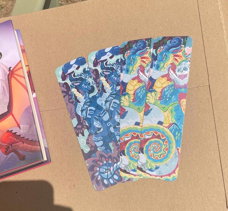 Peut inclure: Quatre marque-pages rectangulaires pr&eacute;sentant une illustration vibrante et multicolore d'un dragon aux &eacute;cailles bleues, jaunes et rouges. Le design comprend des motifs tourbillonnants complexes et un motif de cr&eacute;ature stylis&eacute;, dispos&eacute;s en &eacute;ventail.