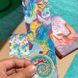 K&ouml;nnte beinhalten: Ein rechteckiges Lesezeichen mit einer farbenfrohen Illustration eines blauen und gr&uuml;nen Drachen, der ein kleines wei&szlig;es Faultier umarmt. Das Design enth&auml;lt leuchtend gelbe, rote und t&uuml;rkise Muster mit einem Spiralmotiv am unteren Ende.