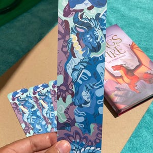 Peut inclure: Un marque-page rectangulaire pr&eacute;sentant une illustration stylis&eacute;e d'un dragon bleu avec des marques blanches et un collier jaune. L'&oelig;uvre utilise des tons de bleu, de violet et de sarcelle. Le marque-page est pr&eacute;sent&eacute; avec un livre Wings of Fire.