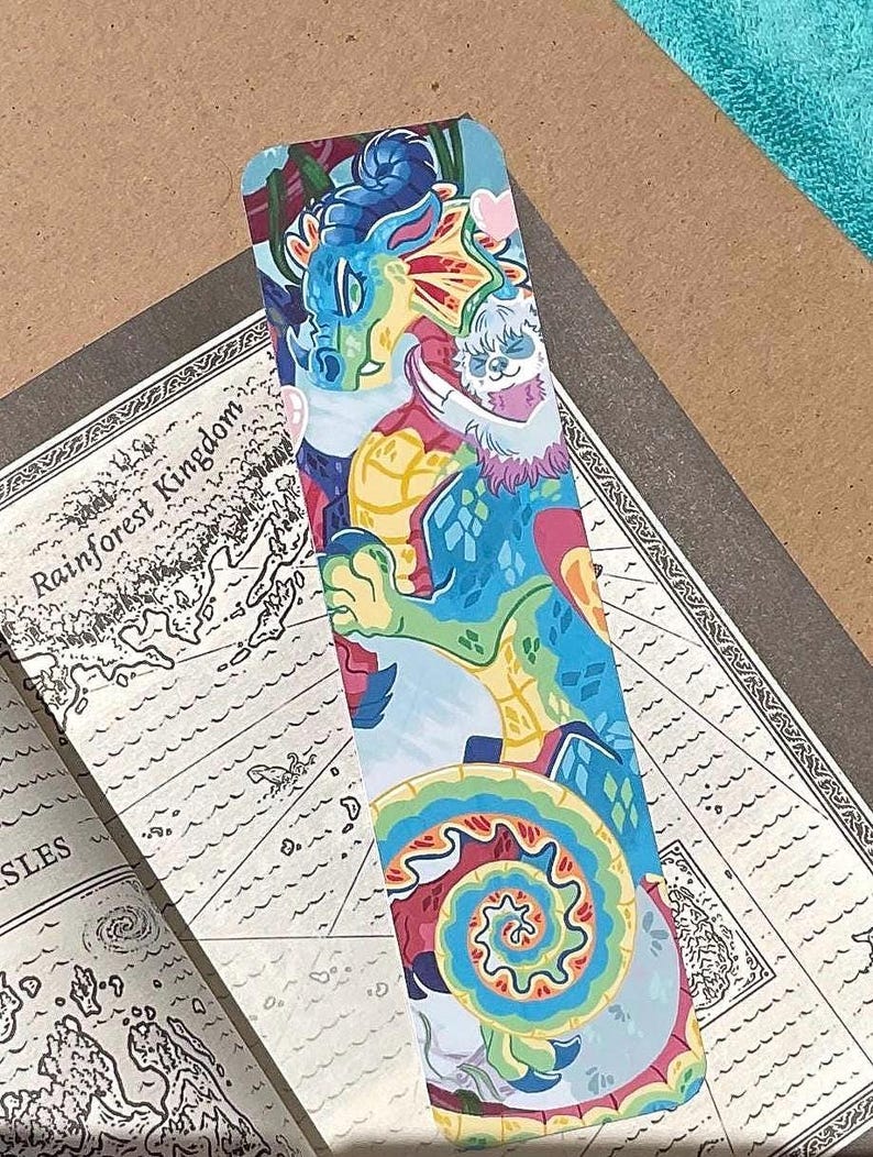K&ouml;nnte beinhalten: Ein rechteckiges Lesezeichen mit einer farbenfrohen Illustration eines blauen und gr&uuml;nen Drachen mit einem kleinen wei&szlig;en, faultierartigen Wesen. Das Kunstwerk enth&auml;lt ein lebhaftes Spiralmuster am unteren Rand und ein kleines rosa Herz.
