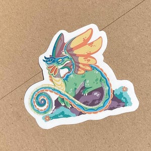 Pu&ograve; includere: Un adesivo fustellato raffigurante un drago verde stilizzato con una lunga coda arricciata, grandi ali gialle e arancioni e accenti blu. Il drago &egrave; appollaiato su una formazione rocciosa viola scuro con piccoli fiori arancioni e fogliame verde acqua.