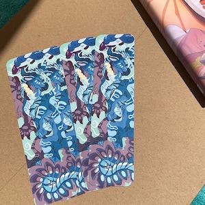 Peut inclure: Trois marque-pages rectangulaires pr&eacute;sentant une illustration r&eacute;p&eacute;t&eacute;e d'une cr&eacute;ature bleue ressemblant &agrave; un tigre avec des marques blanches et des accents jaunes. Le motif int&egrave;gre des volutes violettes et bleu clair, avec un motif floral stylis&eacute; &agrave; la base.