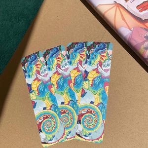 K&ouml;nnte beinhalten: Drei rechteckige Lesezeichen mit einer lebendigen, farbenfrohen Illustration eines Drachen mit blauen, gelben und roten Schuppen. Das Kunstwerk zeigt ein wirbelndes, kreisf&ouml;rmiges Muster am unteren Rand und einen stilisierten Drachenkopf oben.