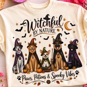 Peut inclure: Un t-shirt crème avec un motif de cinq chiens portant des chapeaux de sorcière et des capes. Le texte indique Witchful BY NATURE et Paws, Potions & Spooky Vibes. Le design inclut des chauves-souris, des étoiles, des feuilles et un chaudron.