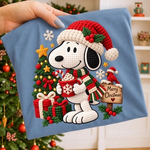 May include: A light blue t-shirt featuring a crochet-style graphic of Snoopy wearing a red Santa hat and a green and red striped scarf. Snoopy holds a red mug with a snowflake design, standing near a decorated Christmas tree and gifts.