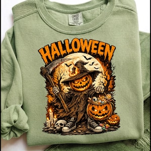 Peut inclure: Un sweat-shirt à col rond vert sauge avec une impression graphique portant le texte HALLOWEEN en orange. Le motif représente une silhouette à tête de citrouille portant un chapeau de sorcière et une faux, à côté d'un seau citrouille rempli de bonbons.