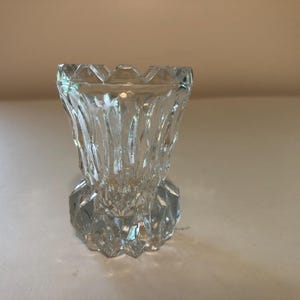 Könnte beinhalten: Ein kleines Gefäß aus klarem Glas mit einem facettierten, geometrischen Design, einem breiten, gewellten Rand und einem dicken, mehrseitigen Boden. Das transparente Glas reflektiert das Licht durch seine kantigen Schliffmuster.