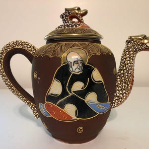 Könnte beinhalten: Eine braune Keramik-Teekanne mit einer handbemalten Figur in einem schwarzen Gewand mit goldenen Akzenten. Der Griff und der Ausguss sind mit weißen, erhabenen Punkten texturiert. Der Deckel ist mit einem goldenen Drachen verziert, und der Körper zeigt goldene Linienmuster sowie kleine Spiralmotive.