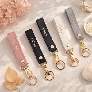Könnte beinhalten: Fünf rechteckige Schlüsselanhänger aus Stoff in Rosa, Schwarz, Schwarz, Weiß und Grau, jeweils mit goldfarbenen Metallbeschlägen, einem Karabinerhaken, einem Ring und einem goldgeprägten Namen: Feerri, Lihua, Chöe, Olauer und Torey.