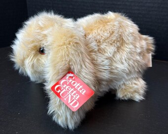 May include: A plush stuffed animal in the shape of a tan rabbit with long, floppy ears and soft, textured fur. The rabbit has dark brown eyes and a red tag attached to its ear that features the white text Gotta Getta GUND printed on it.