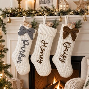 Puede incluir: Tres calcetines navideños de punto trenzado en color crema colgados en fila. Cada calcetín presenta un puño acanalado, un lazo decorativo y un nombre bordado en letra cursiva: Parker en gris, Honey en marrón y Daisy en marrón.
