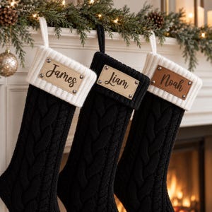 May include: Three cable-knit Christmas stockings featuring a ribbed cuff and a rectangular leather patch secured with four metal rivets. The left stocking is black with a white cuff, the center is black, and the right is black with a white cuff. The patches are engraved with the names James, Liam, and Noah.