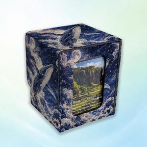 Op de afbeelding: Een vierkante deckbox met een donkerblauwe hemelse en oceanische print met walvissen en beukende golven. Het voorpaneel heeft een rechthoekige uitsparing die een ruilkaart toont met een landschapsillustratie en de tekst Mountain Plains Swamp.