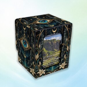 May include: A square deck box featuring a dark, intricate design with gold filigree, crescent moons, and a prominent blue eye. The front panel has a cutout window displaying a landscape card with text including Mountain, Plains, and Swamp.