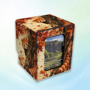 May include: A square card storage box featuring a red panda illustration amidst autumn leaves and tree trunks. The box has a central rectangular cutout displaying a game card with a landscape image and text including Mountain, Plains, and Swamp.