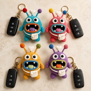 May include: Four crocheted monster keychains in blue, pink, yellow, and purple, each featuring two eyes, a wide mouth with teeth, a small white crossbody bag, and two antennae topped with colourful spheres and a central white heart shape.